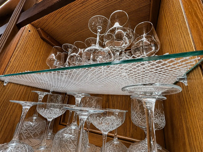 A collection of clear cut-crystal wine glasses and coupe glasses displayed on toughened glass shelves cut to size inside a wooden cabinet.